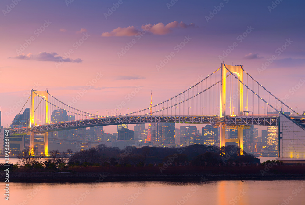 Obraz premium Rainbow Bridge illuminated at twilight with Tokyo skyline and Tokyo Tower in the background, reflecting on calm waterfront under a colorful pastel sunset sky in Tokyo, Japan.