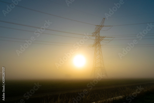 Фотография Sunrise behind foggy electricity pylon over rural field with high voltage power
