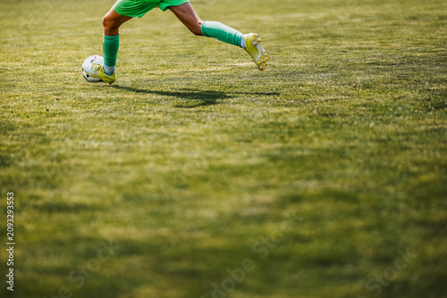 Cropped view of a soccer player in a green uniform running with the ball on a sunlit grass field, with open copy space in the foreground.