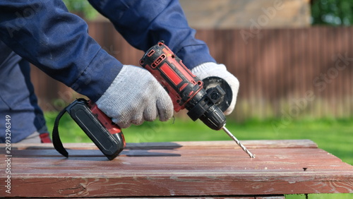 Bild auf Leinwand A close-up of a worker in a blue jacket and protective gloves drilling a hole in a wooden table outdoors with a cordless impact drill
