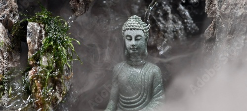 Fotografi A small stone Buddha statue in meditative posture surrounded by moss‑covered rocks and mist