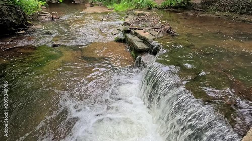 Water cascades down a woodland waterfall into a gentle stream as the camera slowly pulls back in slow motion, revealing the tranquil forest and peaceful natural surroundings.