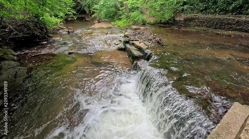 Water cascades down a small woodland waterfall into a gently flowing stream. Filmed in a static slow-motion shot, the scene highlights the peaceful movement of water through a tranquil forest setting.