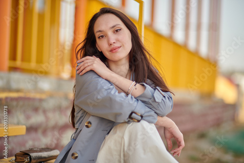 A young woman is sitting on the yellow steps. Her gray coat hangs loosely over her white skirt and dark shoes.