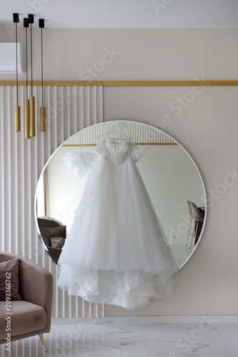 White wedding dress hanging on a round mirror in a modern bright interior, creating an elegant bridal preparation atmosphere.
