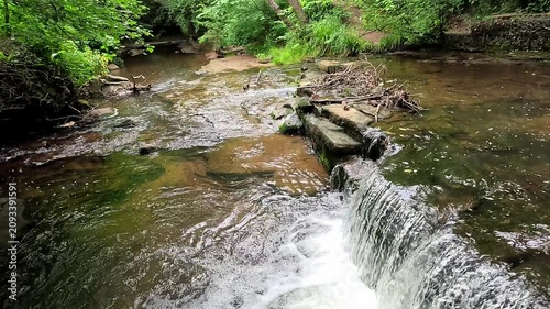 Water cascades over a small woodland waterfall into a gentle stream as the camera slowly pans in super slow motion, revealing every ripple and splash in the tranquil forest setting.