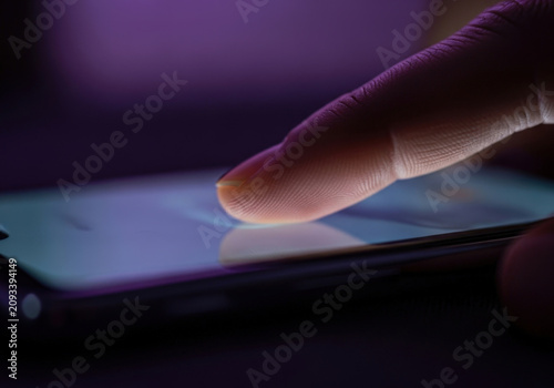 A close up macro shot of a person's finger touching a glowing smartphone screen in low light with purple hues.