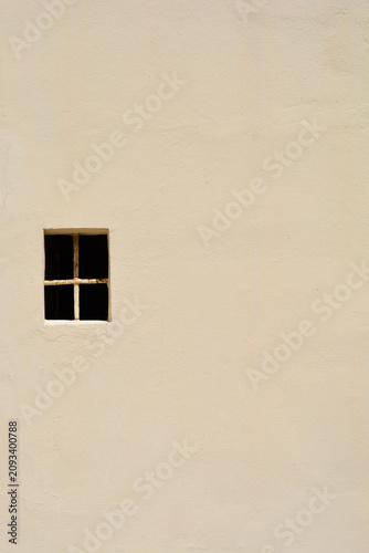 Obraz na plátně A tiny dark square window with a black crossbar structure is positioned on a vast textured beige plaster building facade