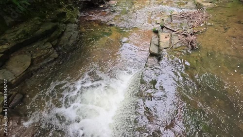 A high-up view captures water cascading over a woodland waterfall in slow motion. The static shot highlights the flowing stream, ripples, rocks, and peaceful forest surroundings.