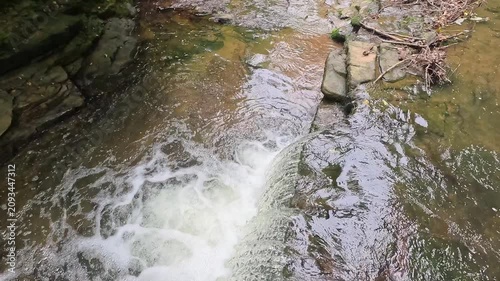 A high-up view follows water cascading over a woodland waterfall in slow motion as the camera pulls back, revealing the winding stream, mossy rocks, and tranquil forest landscape.