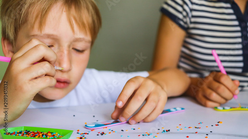 sister and brother are making crafts from diamond mosaics in the form of bracelets on a table in the room close-up