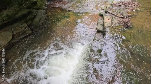 A high-up view moves closer in slow motion towards water cascading over a woodland waterfall, revealing the flowing stream, ripples, rocks, and serene forest scenery below.