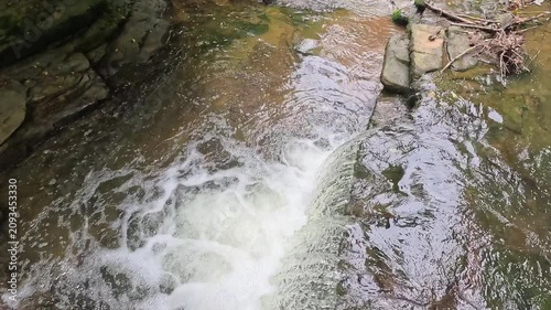 A slow-motion panning shot from above reveals water cascading over a woodland waterfall, capturing the flowing stream, textured rocks, ripples, and peaceful forest landscape.