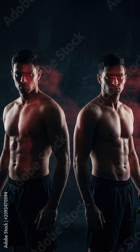 Dual Strength: Two muscular men with sculpted physiques stand side by side, their powerful bodies illuminated by dramatic red lighting against a dark, smoky background.