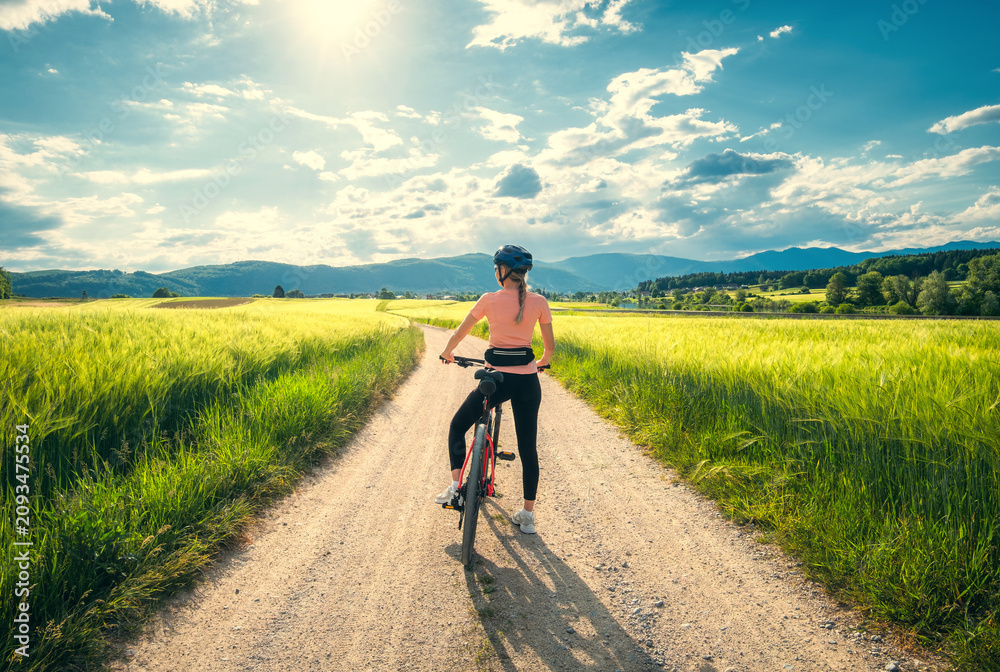 Obraz premium Woman riding mountain bike on dirt road through green wheat fields at sunset in Slovenia. Female cyclist cycling in countryside landscape and sunny sky. Summer outdoor activity, fitness. Girl on bike