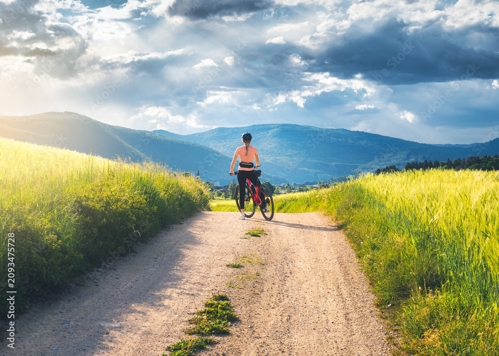 Obraz premium Female cyclist with mountain bike standing on dirt road in Slovenia countryside in summer. Woman looking at beautiful valley and mountains under dramatic stormy sky. Summer outdoor activity