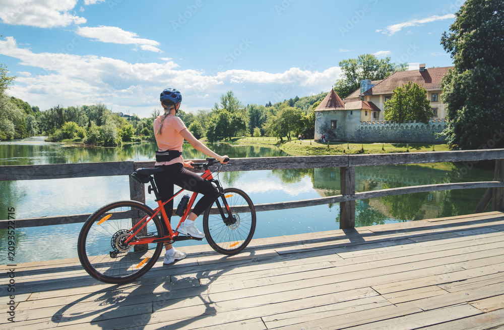 Fototapeta premium Female cyclist with mountain bike standing on wooden bridge in Slovenia. Woman looking at historic Otocec Castle on Krka river under blue sky. Summer cycling trip, travel destination. Girl on bike
