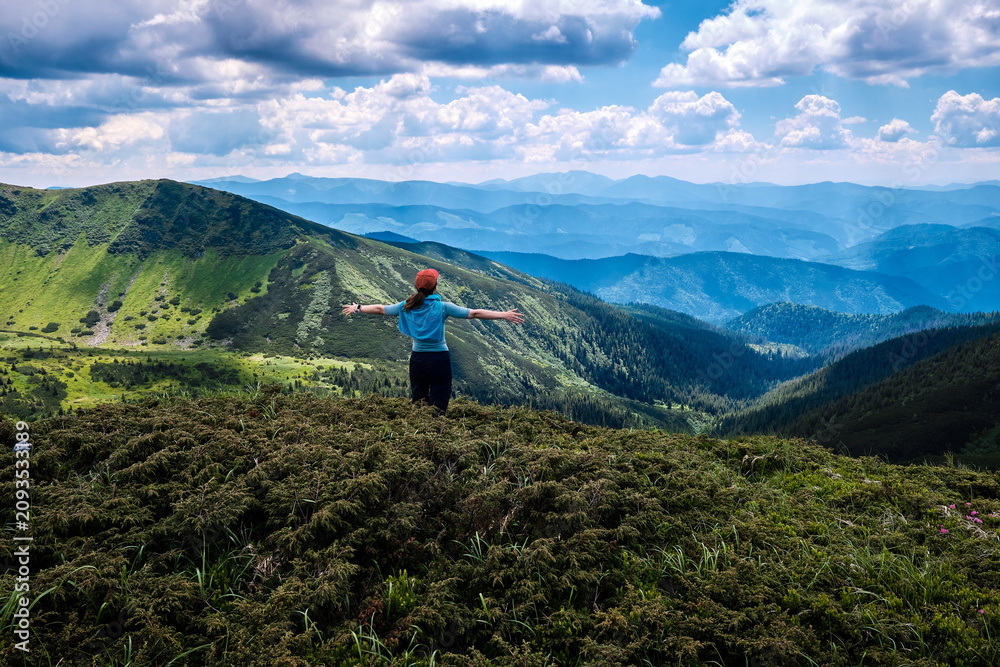 Obraz premium female hiker standing on a lush green mountain summit with her arms spread wide, embracing the freedom and the breathtaking panorama of layered mountain ridges and valleys under a cloudy sky