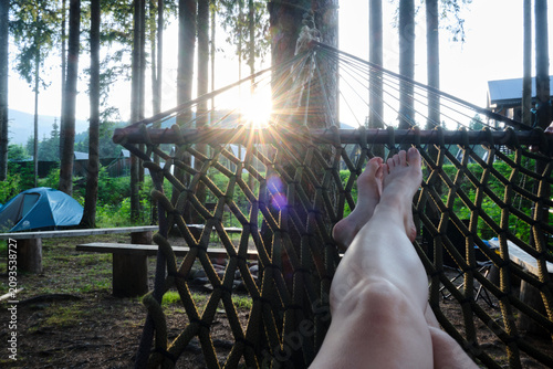 A horizontal point-of-view shot of a person's bare legs and feet resting in a green net hammock at a campground surrounded by tall pine trees and tents.