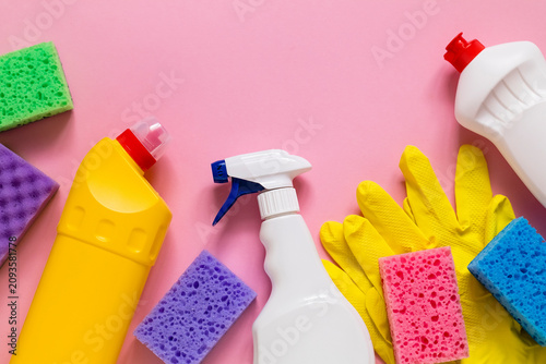 Top view of colorful household cleaning supplies including spray bottles, sponges, rubber gloves, and detergent arranged on a pink background with copy space.