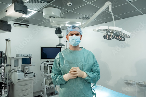 Man surgeon stands in operating room with mask and gloves under surgical lights. Focused medic clasps hands waiting calmly before procedure