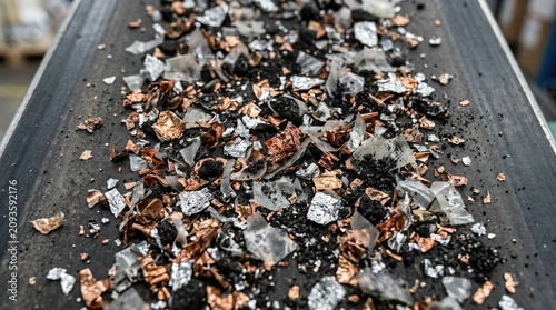 Recycled battery shreds, crushed copper, aluminum foil pieces, dark lithium black mass powder, plastic fragments, industrial waste moving on a conveyor belt, electronic recovery process.