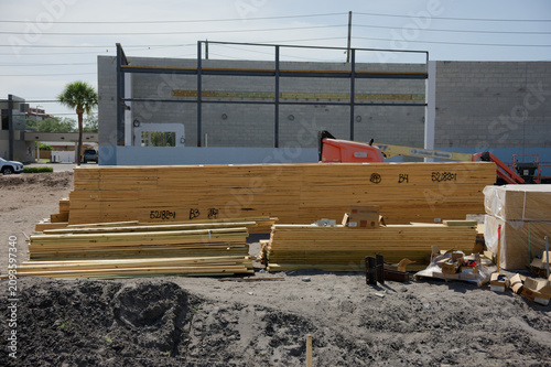 Obraz na płótnie Stacked lumber and packed building materials sit on a dusty outdoor construction site in front of a partially built steel and concrete frame, with construction equipment visible in the background