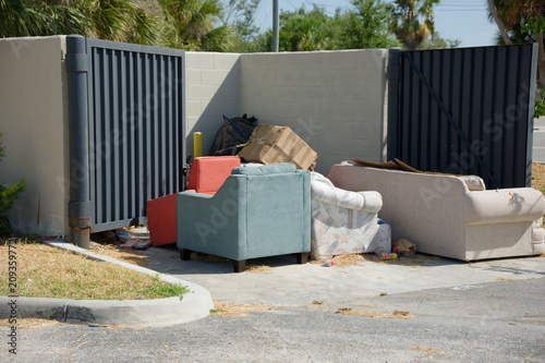 Discarded sofas and armchairs sit beside a metal dumpster enclosure at a residential curb, surrounded by cardboard and trash, suggesting illegal dumping, neglect and bulky furniture disposal on a sunn