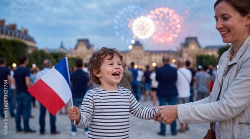 A laughing toddler waves a French flag while holding his mother's hand under distant Bastille Day fireworks for candid family stock photography