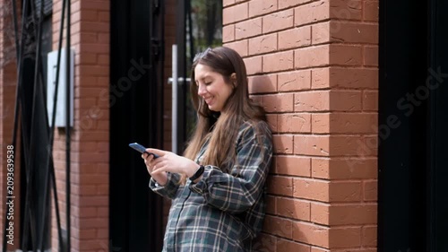 Pregnant woman leaning beside red brick building entrance while checking smartphone and smiling in relaxed urban setting with soft natural daylight and modern street architecture