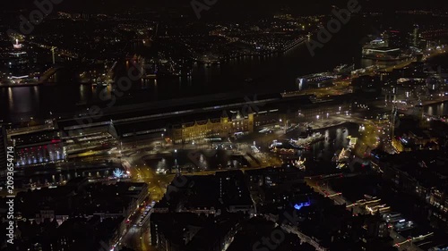 Aerial night view of Amsterdam Central Station, Netherlands, with the illuminated railway terminal, ferry docks on the IJ river, canal bridges, holiday lights and traffic in the city center.