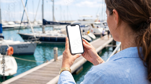 Hand holding smartphone with blank white screen in yacht marina, woman using mobile app for boat travel navigation mockup, luxury summer holiday, digital nomad, sea vacation life.
