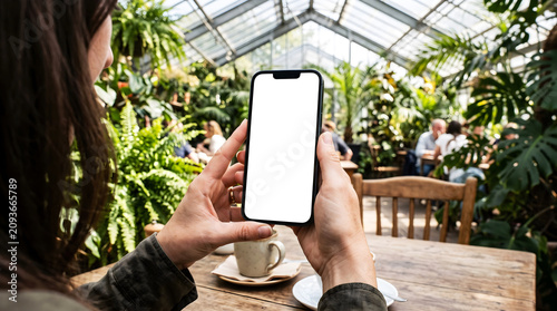 Hand holding smartphone with blank white screen in botanical greenhouse cafe, woman using mobile device for plant store app mockup, gardening research, nature lover interior lifestyle.
