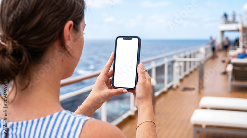 Hand holding smartphone with blank white screen on cruise ship deck, woman using mobile app for travel itinerary mockup, ocean vacation, luxury sea holiday, digital nomad life concept.
