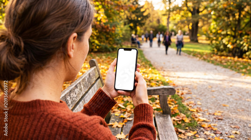 Hand holding smartphone with blank white screen on park bench, woman using mobile app for autumn nature walk navigation, fall seasonal lifestyle, digital content mockup, park path.
