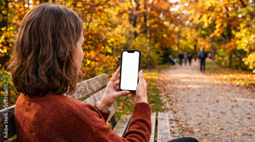 Hand holding mobile phone with blank white screen while sitting on bench in autumn park, woman using smartphone for travel app mockup, nature lifestyle, fall season digital content.
