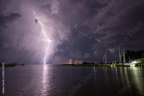 Storm clouds gather over the water with lightning striking in the distance while boats are docked along the shore