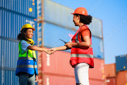 Unity and teamwork concept. Hand shake African woman logistics workers in hardhat helmet and high visibility safety gear at shipping container yard