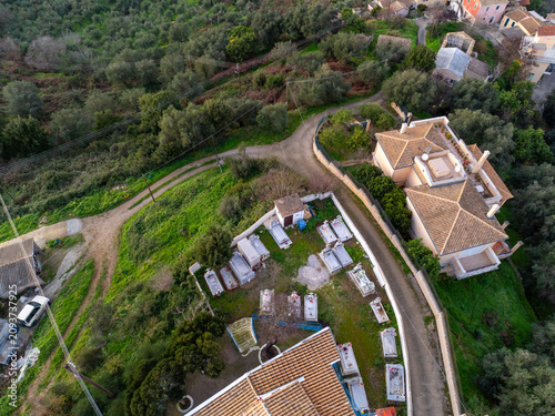 Aerial view of a traditional Greek Orthodox cemetery with white marble tombs and a tiled roof house nestled in a lush green olive grove in Corfu