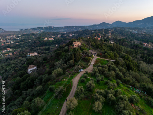 Aerial view of a winding road on a hill ridge with houses and olive trees during sunset in Corfu Greece overlooking the distant sea