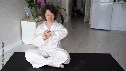 Static Shot Of Woman In White Meditation Clothes Checking Wristwatch Then Holding Prayer Pose On Yoga Mat In Home Interior During Quiet Morning Routine.