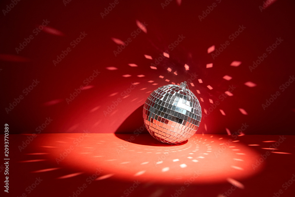 Fototapeta premium Shiny disco ball, also known as mirror ball, on a red surface with scattered light reflections. Disco ball decoration with light reflections on vibrant red background
