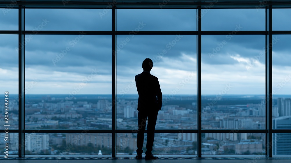 Fototapeta premium Silhouette of a businessman in a suit standing in a modern office, looking out at a cityscape through a large window