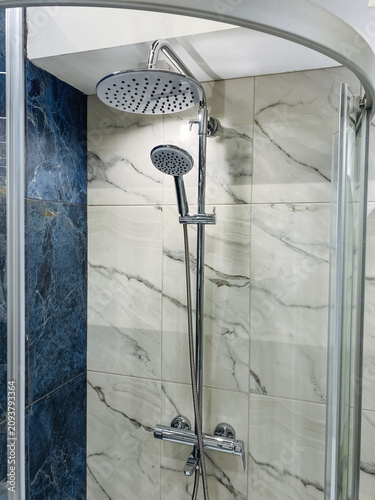 Modern bathroom shower with large round rainfall head, handheld wand, chrome rail, marble tile walls, glass enclosure and polished fixtures in bright clean interior