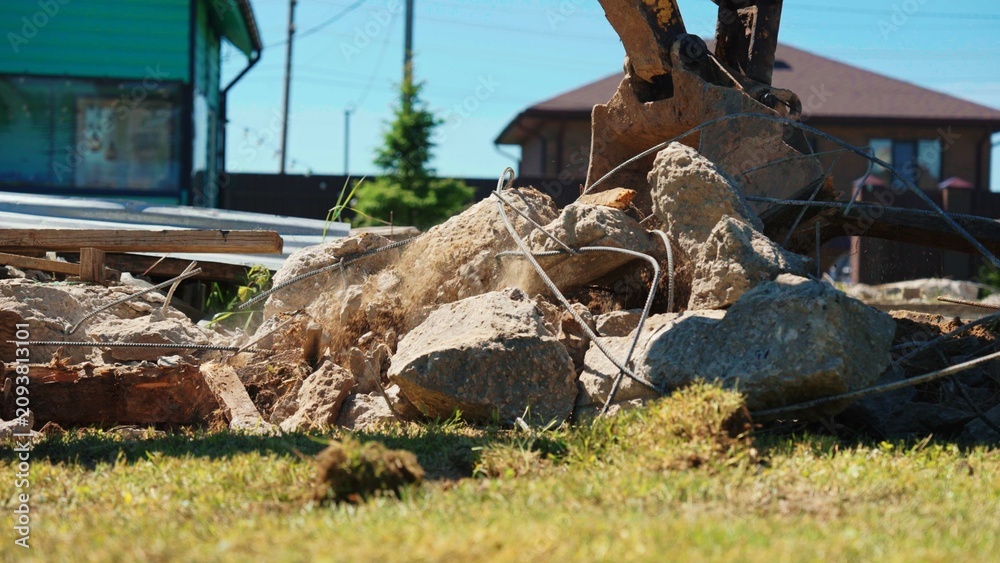 Fototapeta premium Excavator bucket actively breaking reinforced concrete and rebar from a demolished structure, creating rubble on a sunny construction site with a residential background