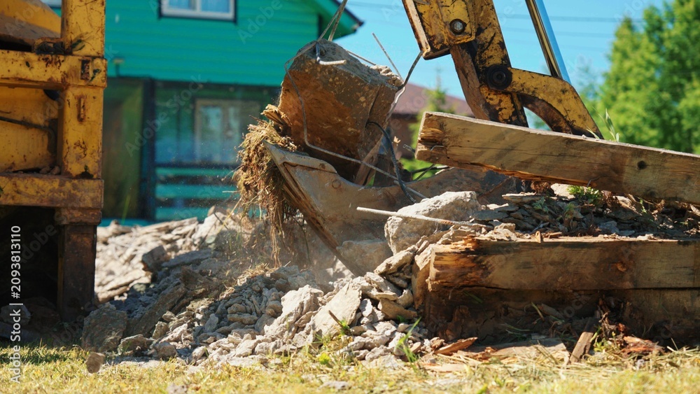 Fototapeta premium Low-angle view of a backhoe bucket holding a reinforced concrete block above demolition rubble, with bent rebar, hanging roots and broken timber at a residential site.