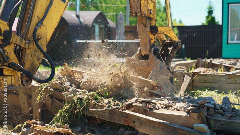 Fototapeta premium Yellow excavator bucket breaking up rubble and wooden debris on a sunny day, clearing the ground for new development and construction work in a suburban area