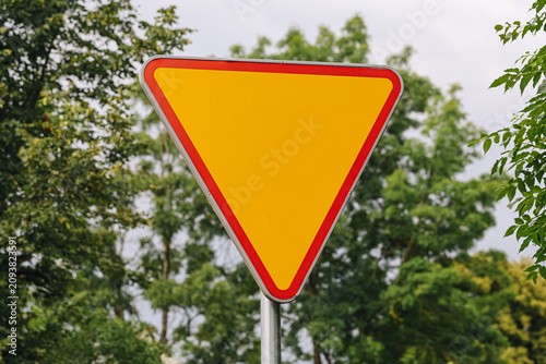 Blank yield sign stands outdoors. Yellow triangle has red border. Green trees create natural background. Empty road sign allows customization. Traffic concept supports safety designs.