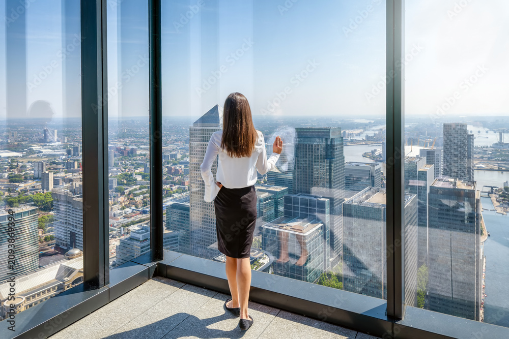 Obraz premium A business woman in corporate outfit looks at the skyline of the financial district Canary Wharf in London from her office window