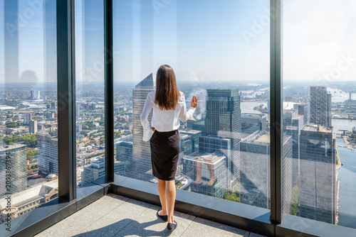 A business woman in corporate outfit looks at the skyline of the financial district Canary Wharf in London from her office window